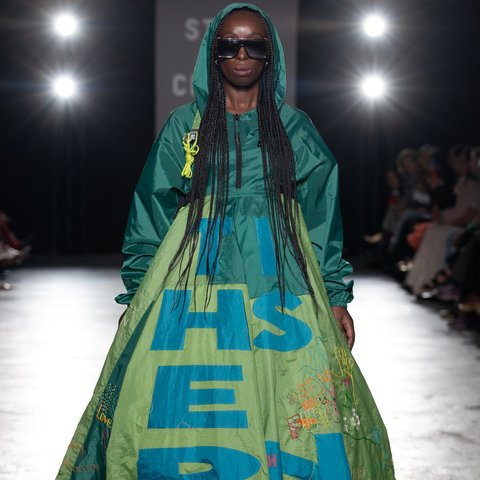 A model walks the runway in a green hooded dress with oversized text and embroidered details, accessorised with large black sunglasses. The dress features bold blue lettering and colourful stitched embellishments.