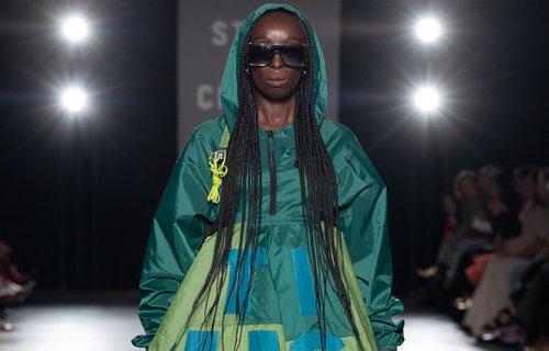 A model walks the runway in a green hooded dress with oversized text and embroidered details, accessorised with large black sunglasses. The dress features bold blue lettering and colourful stitched embellishments.
