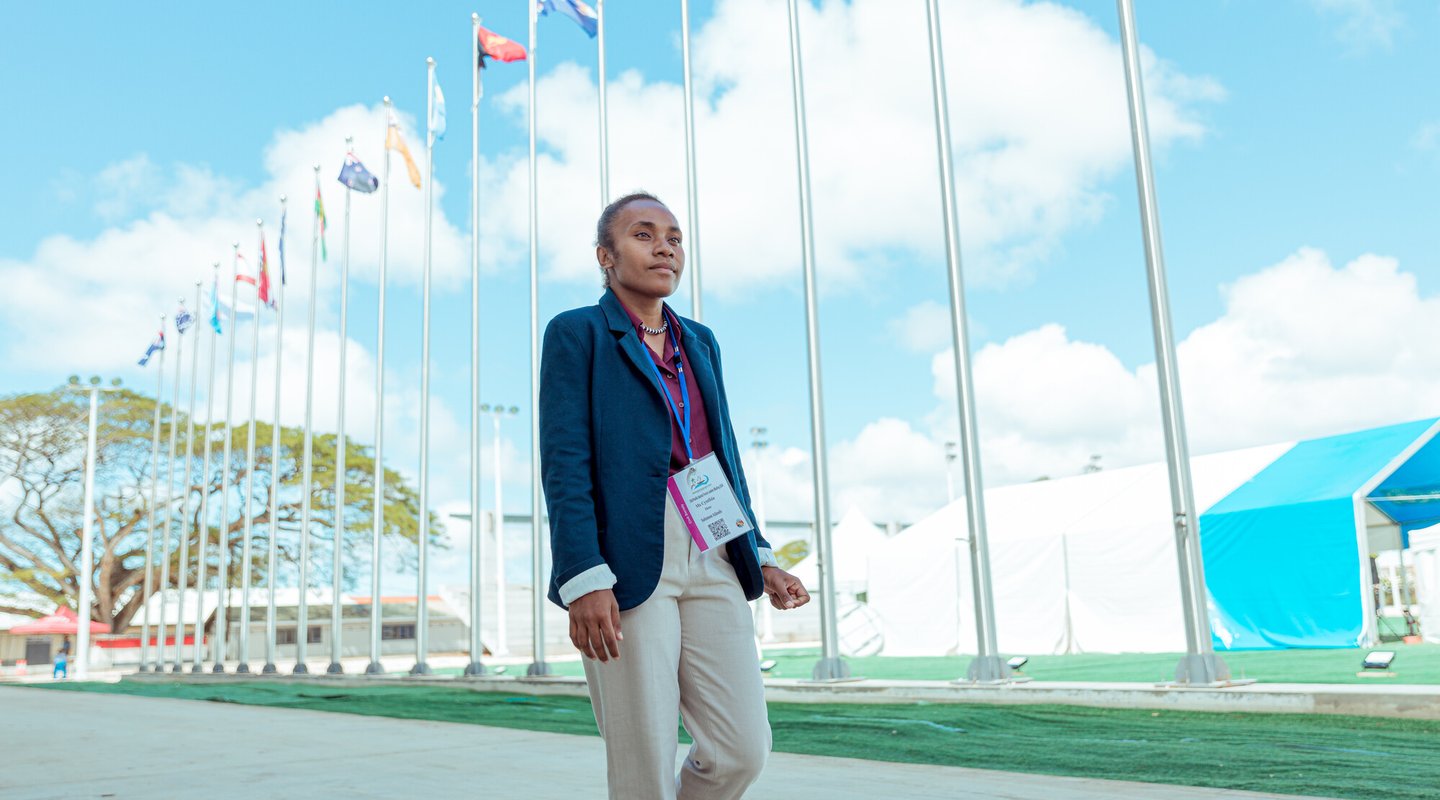 Cynthia stands wearing a lanyard outside a conference hall with flags behind her.