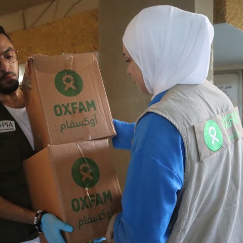 A woman aid worker in Lebanon wearing a white hijab and Oxfam jacket hands a box with Oxfam written on it to a male aid worker wearing a jacket with Utopia, an Oxfam partner name, written on it.