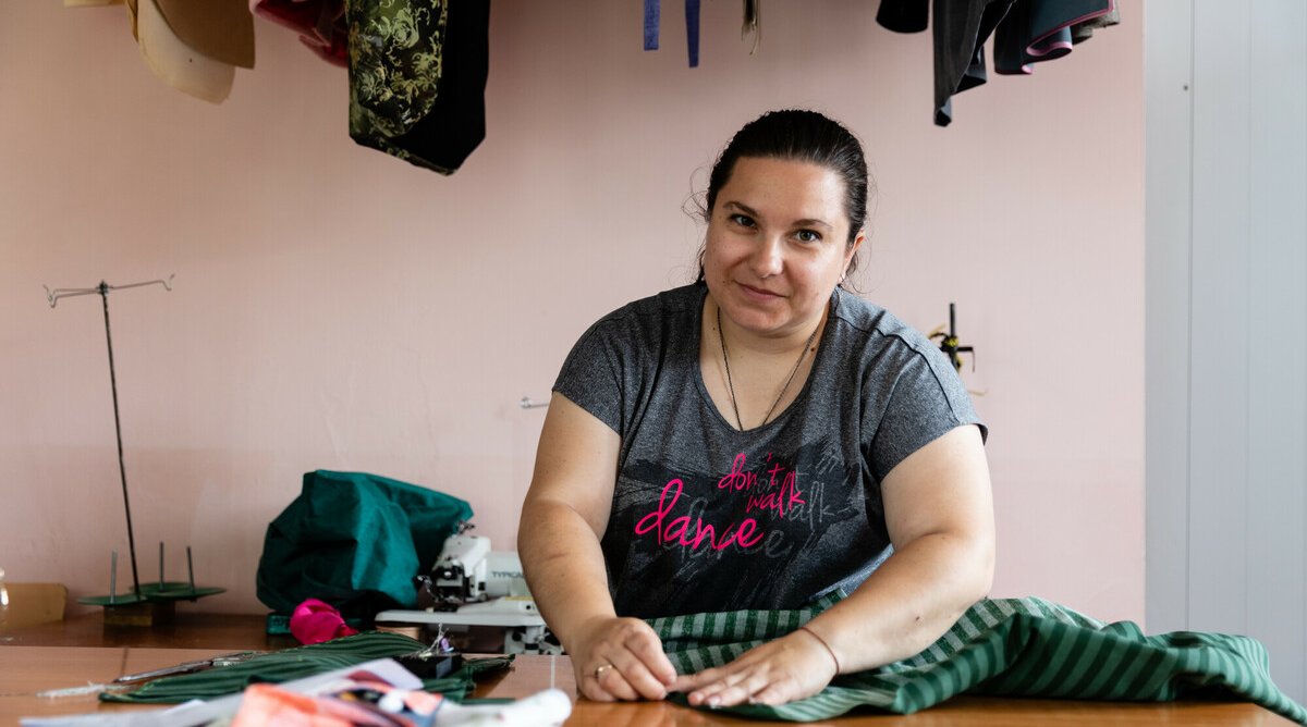 A person sits at a sewing table working with striped fabric, surrounded by sewing materials and garments hanging overhead.