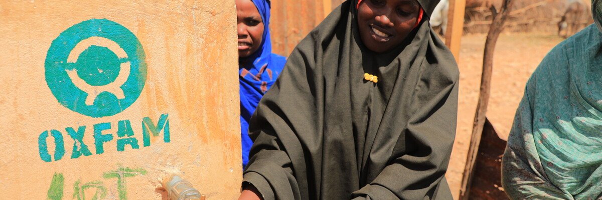 People collect water from an Oxfam-constructed water point, filling a yellow jerrycan in a dry, outdoor setting. The Oxfam logo is painted on the water tank beside them.