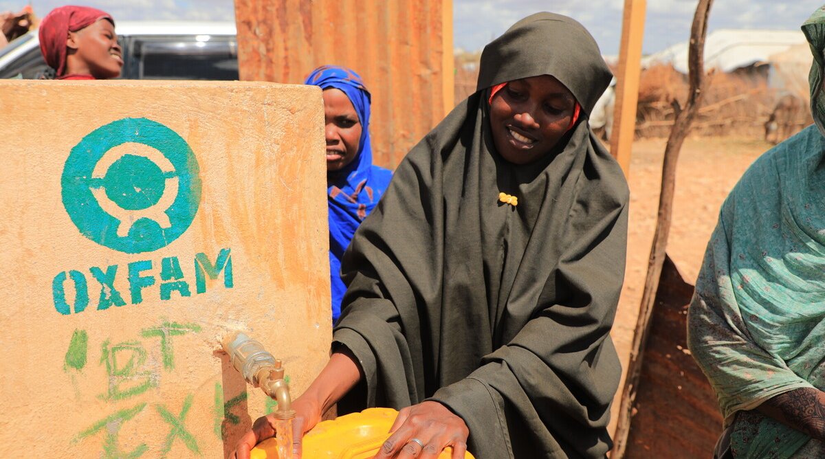 People collect water from an Oxfam-constructed water point, filling a yellow jerrycan in a dry, outdoor setting. The Oxfam logo is painted on the water tank beside them.