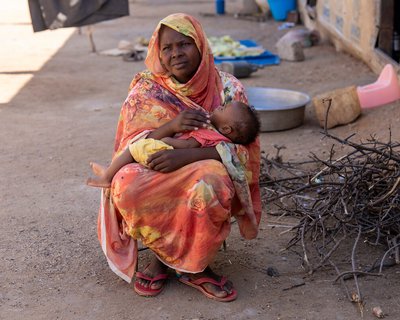 A woman sits outside holding a young child in her arms, wrapped in bright patterned fabric. Nearby are cooking pots, collected firewood, and household items on the ground.