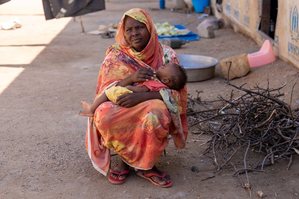 A woman sits outside holding a young child in her arms, wrapped in bright patterned fabric. Nearby are cooking pots, collected firewood, and household items on the ground.