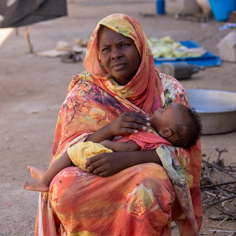 A woman sits outside holding a young child in her arms, wrapped in bright patterned fabric. Nearby are cooking pots, collected firewood, and household items on the ground.