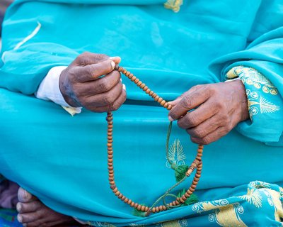 A close up of the hands of a woman sitting cross-legged. She is holding a string of prayer beads/