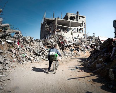 A person pushes a bicycle through a street filled with rubble and destroyed buildings in a heavily damaged urban area.