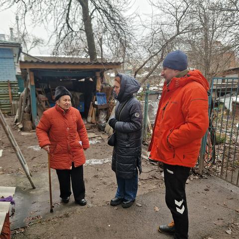 Hanna is standing with a walking stick outside her home that has been damaged by shelling. It is winter and they are all wearing big coats and hats. Hanna is talking to Anika who is visiting to see the newly installed windows.