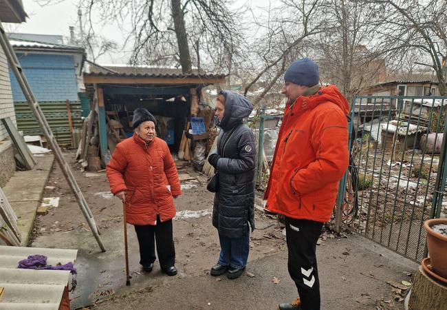 Hanna is standing with a walking stick outside her home that has been damaged by shelling. It is winter and they are all wearing big coats and hats. Hanna is talking to Anika who is visiting to see the newly installed windows.