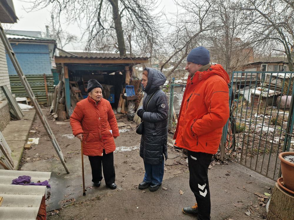 Hanna is standing with a walking stick outside her home that has been damaged by shelling. It is winter and they are all wearing big coats and hats. Hanna is talking to Anika who is visiting to see the newly installed windows.