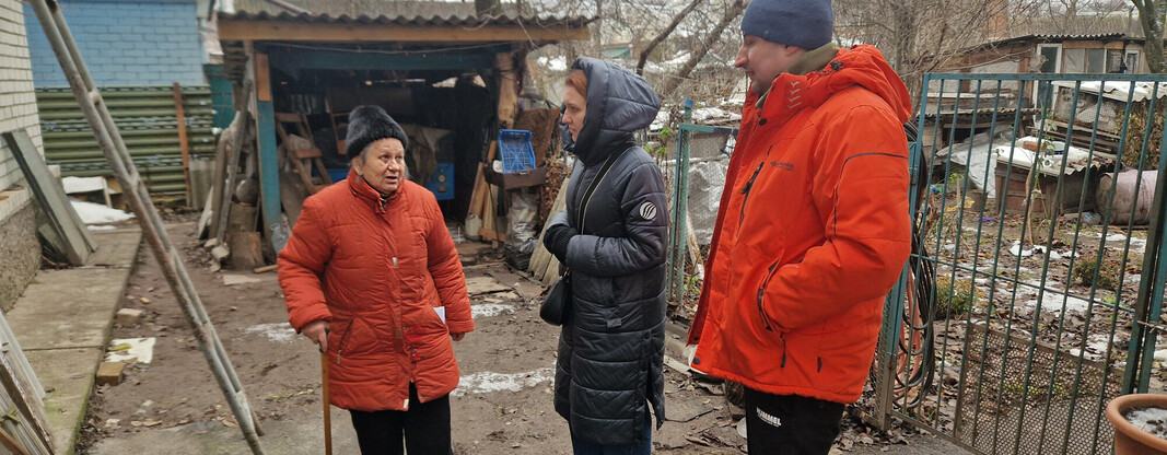 Hanna is standing with a walking stick outside her home that has been damaged by shelling. It is winter and they are all wearing big coats and hats. Hanna is talking to Anika who is visiting to see the newly installed windows.