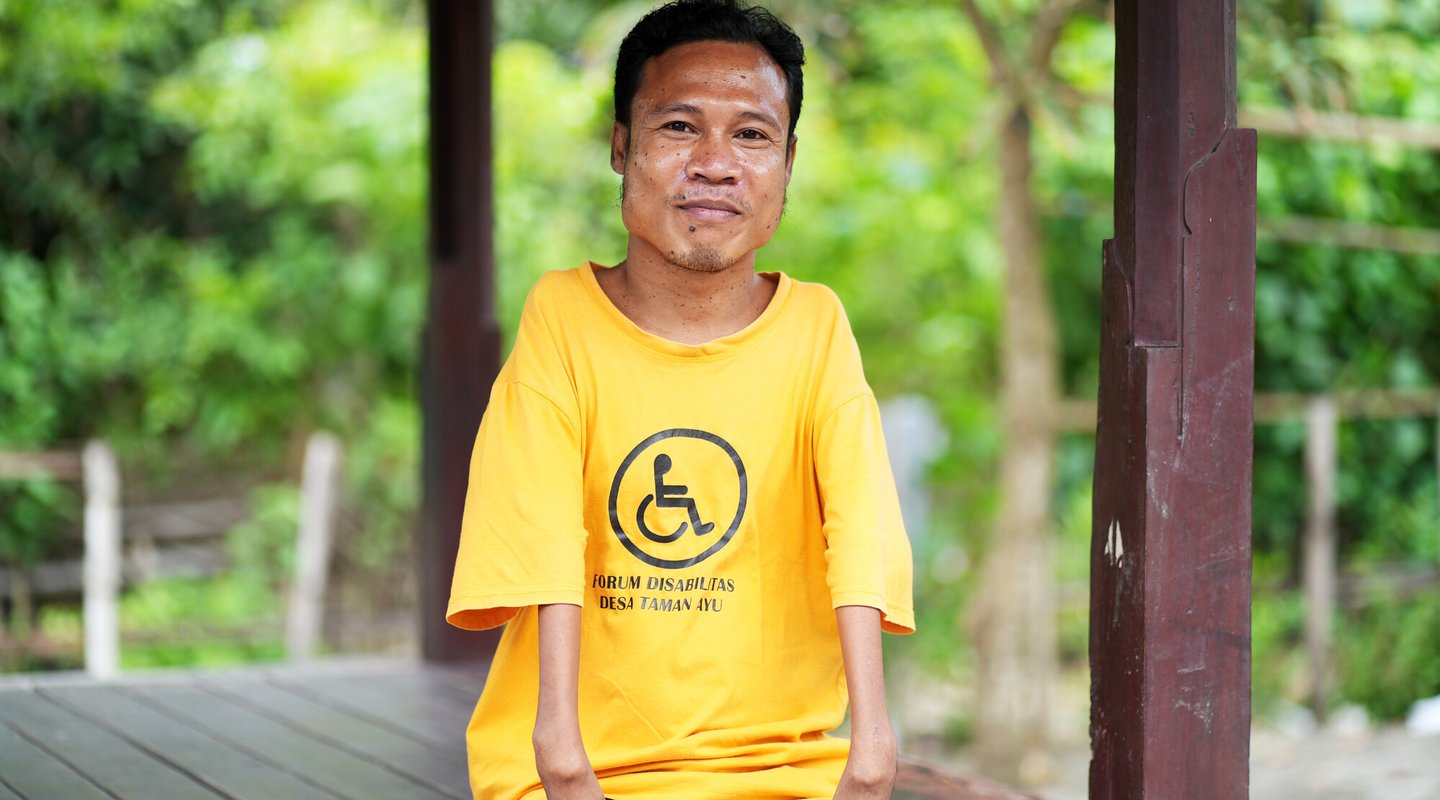 A man sitting outside, wearing a t-shirt saying “forum disabilitas desa Taman Ayu” [translation: Taman Ayu Village Disability Forum]