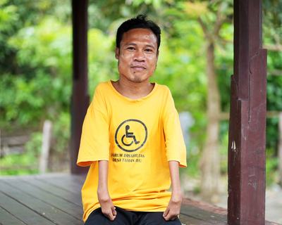 A man sitting outside, wearing a t-shirt saying “forum disabilitas desa Taman Ayu” [translation: Taman Ayu Village Disability Forum]