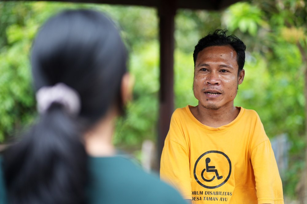 A man sitting outside, speaking to a woman. He's wearing a t-shirt that says "forum disabilitas desa Taman Ayu” [translation: Taman Ayu Village Disability Forum]