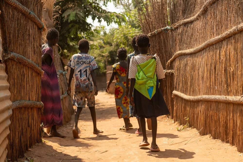 A group of children walk barefoot down a sunlit path between traditional reed fences in a rural village. One child carries a bright green backpack.