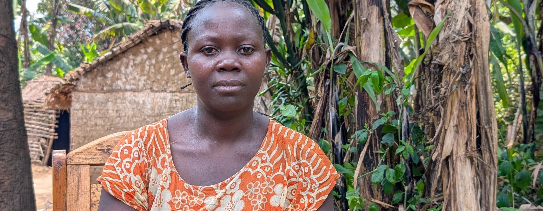 Kahindo wearing an orange dress with white floral patterns sits on a wooden chair outdoors, surrounded by lush green vegetation and banana trees, with a thatched hut visible in the background.