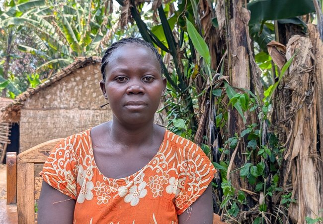 Kahindo wearing an orange dress with white floral patterns sits on a wooden chair outdoors, surrounded by lush green vegetation and banana trees, with a thatched hut visible in the background.