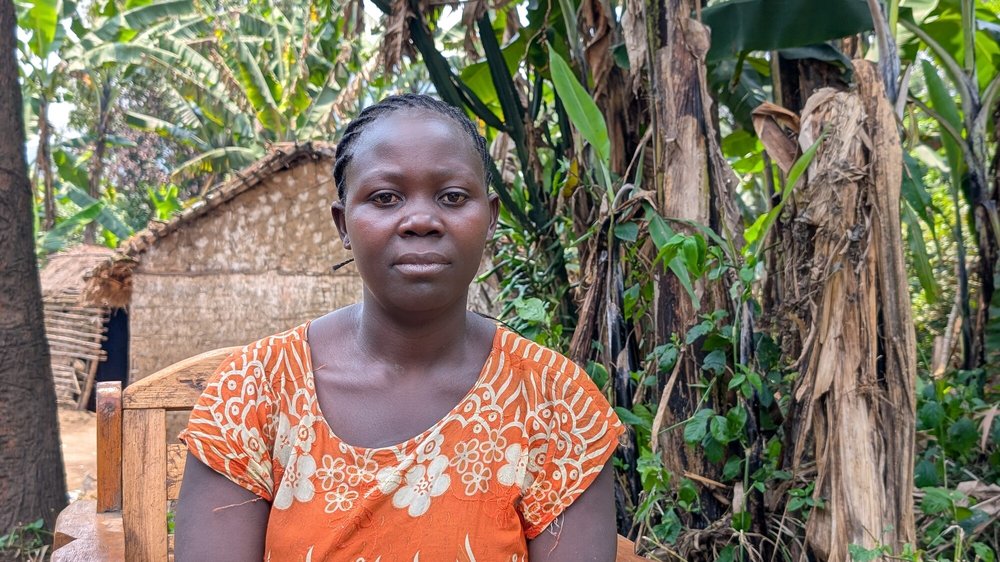 Kahindo wearing an orange dress with white floral patterns sits on a wooden chair outdoors, surrounded by lush green vegetation and banana trees, with a thatched hut visible in the background.