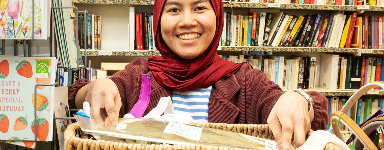 Ria, a volunteer at Brighton London Road Oxfam shop, stands smiling behind a rack of clothes.