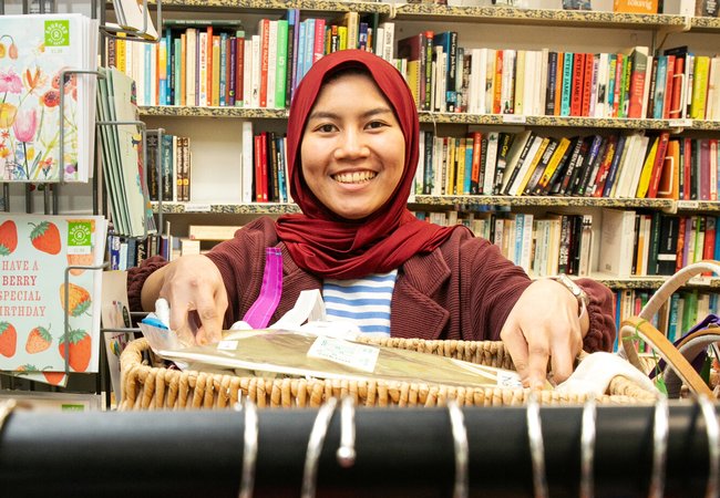 Ria, a volunteer at Brighton London Road Oxfam shop, stands smiling behind a rack of clothes.