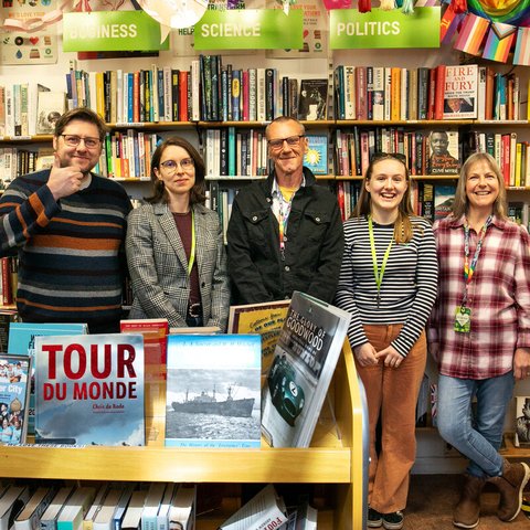 Group of people standing in a bookshop, they are all smiling and one is giving the thumbs up sign.
