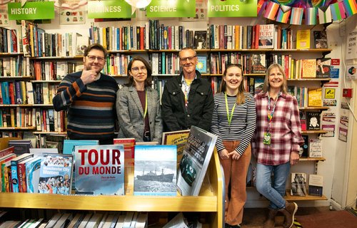 Group of people standing in a bookshop, they are all smiling and one is giving the thumbs up sign.