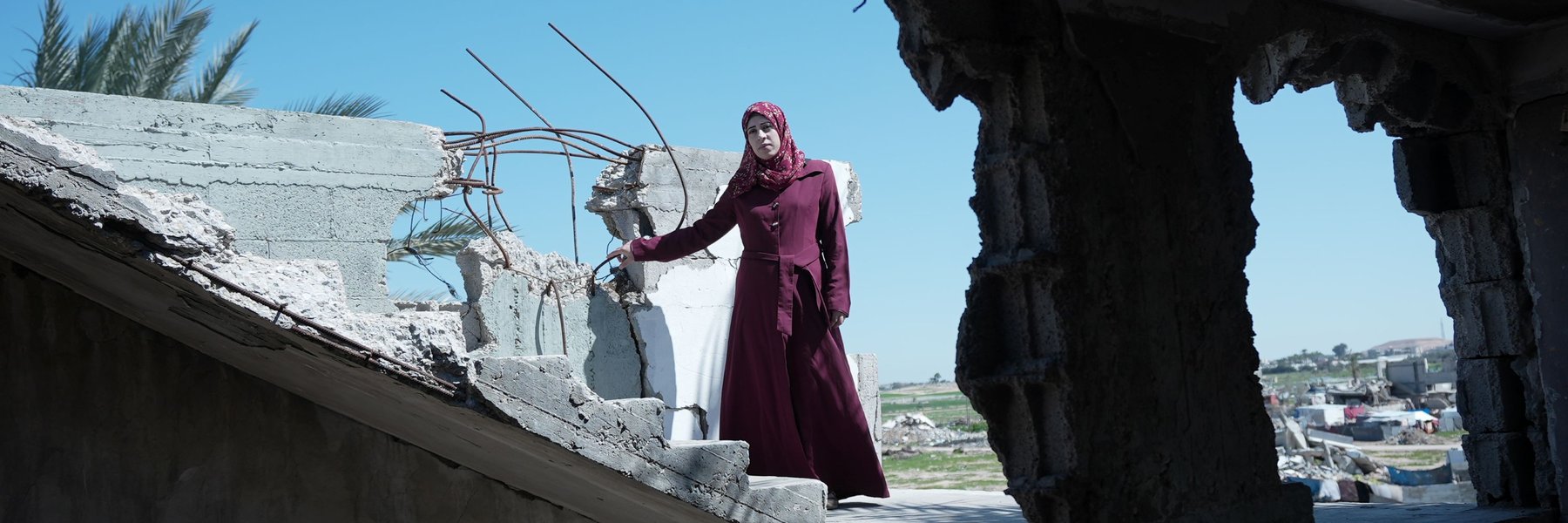A woman in a maroon dress stands among the rubble of a destroyed building under a clear blue sky. She is Suhad, a mother of six in Gaza, facing the hardships of war and displacement. She received maternal care through the Power to Choose Program.