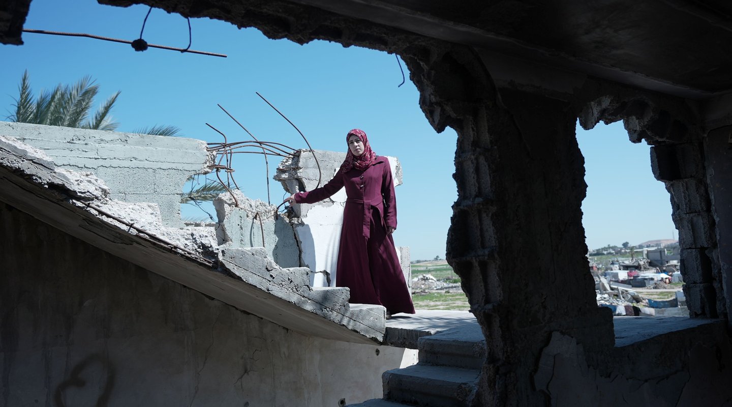 A woman in a maroon dress stands among the rubble of a destroyed building under a clear blue sky. She is Suhad, a mother of six in Gaza, facing the hardships of war and displacement. She received maternal care through the Power to Choose Program.