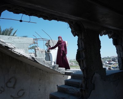 A woman in a maroon dress stands among the rubble of a destroyed building under a clear blue sky. She is Suhad, a mother of six in Gaza, facing the hardships of war and displacement. She received maternal care through the Power to Choose Program.