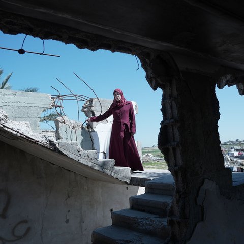 A woman in a maroon dress stands among the rubble of a destroyed building under a clear blue sky. She is Suhad, a mother of six in Gaza, facing the hardships of war and displacement. She received maternal care through the Power to Choose Program.