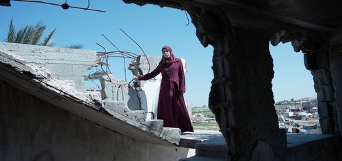 A woman in a maroon dress stands among the rubble of a destroyed building under a clear blue sky. She is Suhad, a mother of six in Gaza, facing the hardships of war and displacement. She received maternal care through the Power to Choose Program.