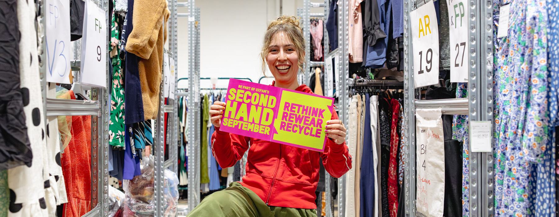 Amy modelling second hand clothes and holding Second Hand September poster in between rails in Oxfam's warehouse