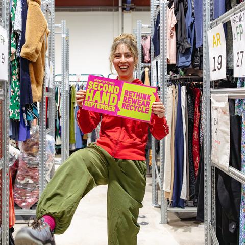 Amy modelling second hand clothes and holding Second Hand September poster in between rails in Oxfam's warehouse