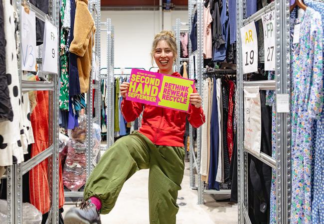 Amy modelling second hand clothes and holding Second Hand September poster in between rails in Oxfam's warehouse