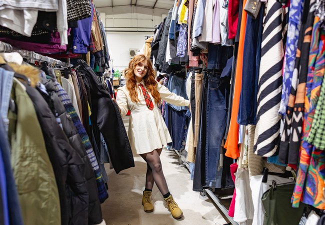 A person stands in the aisle of a secondhand clothing store, surrounded by racks filled with assorted garments like jackets, jeans, shirts, and colorful dresses. The individual is wearing a cream-colored dress with a check tie, black tights, and tan boots