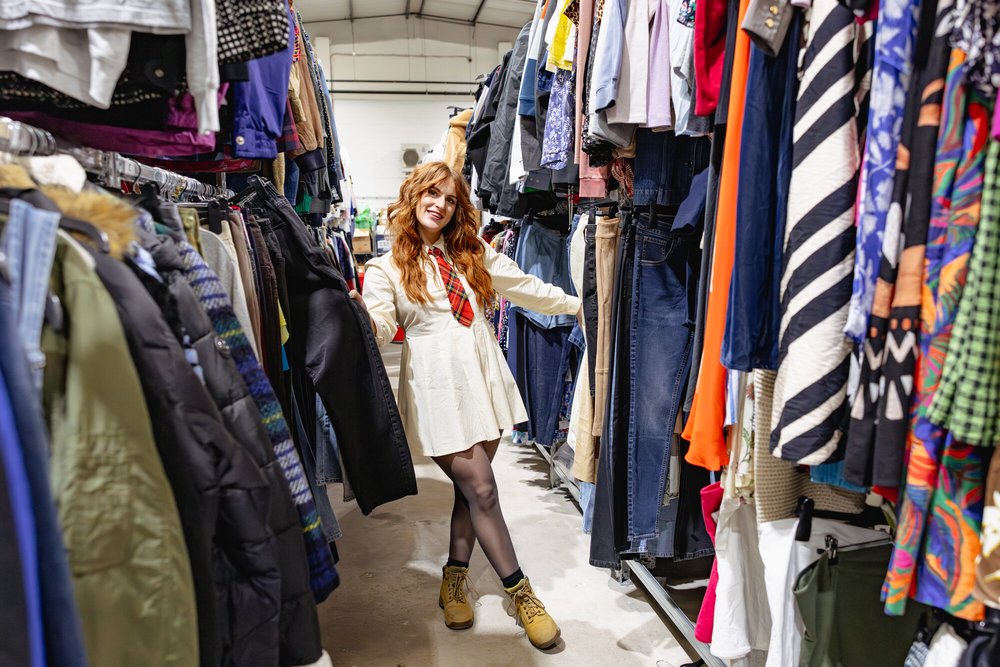A person stands in the aisle of a secondhand clothing store, surrounded by racks filled with assorted garments like jackets, jeans, shirts, and colorful dresses. The individual is wearing a cream-colored dress with a check tie, black tights, and tan boots
