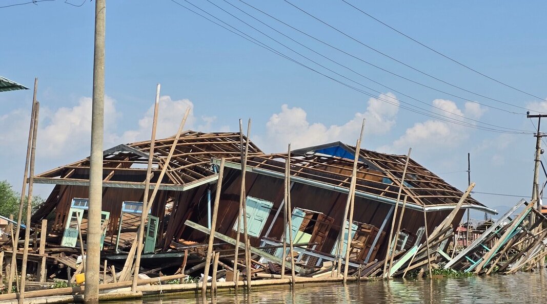 A wooden house has collapsed and partially submerged in floodwater, with leaning utility poles and debris visible under a clear blue sky.