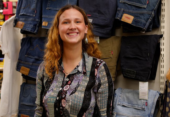 A smiling Oxfam volunteer with red hair and a nose ring stands in front of a display of pairs of jeans in an Oxfam shop.