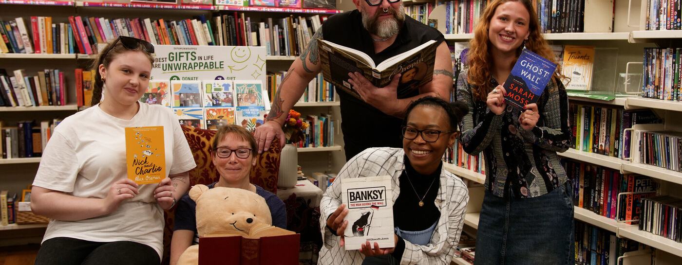A group of smiling people in an Oxfam bookshop pose among bookshelves, each holding a book while surrounded by shelves filled with second-hand books.