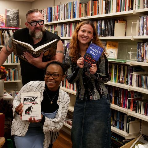 A group of smiling people in an Oxfam bookshop pose among bookshelves, each holding a book while surrounded by shelves filled with second-hand books.