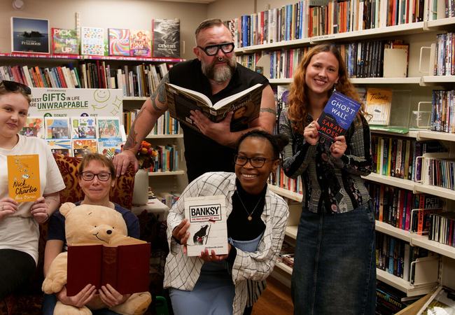 A group of smiling people in an Oxfam bookshop pose among bookshelves, each holding a book while surrounded by shelves filled with second-hand books.