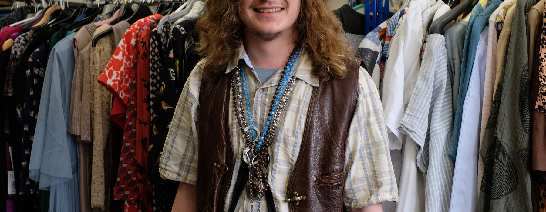 A smiling, long-haired man in a leather waistcoat stands in front of a rack of donated clothes in an Oxfam shop.