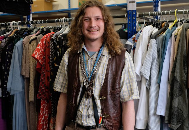 A smiling, long-haired man in a leather waistcoat stands in front of a rack of donated clothes in an Oxfam shop.