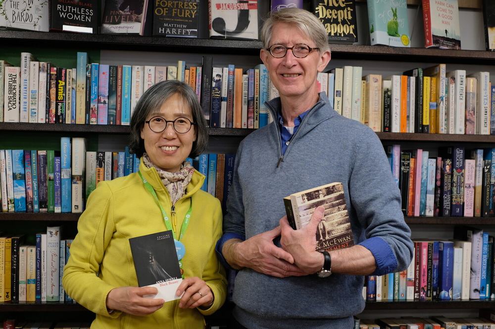 Two smiling people standing in front of bookshelves in a bookshop, each holding a book. Rows of fiction and non-fiction books fill the shelves behind them.