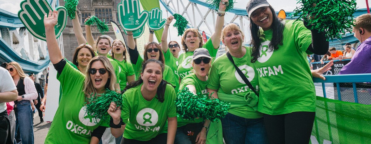 Oxfam volunteers, wearing green Oxfam logo t-shirts, pose for a group photo on Tower Bridge at the London Marathon 2025. They wave pom-poms and smile at the camera.