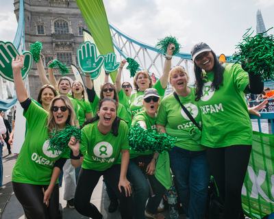 Oxfam volunteers, wearing green Oxfam logo t-shirts, pose for a group photo on Tower Bridge at the London Marathon 2025. They wave pom-poms and smile at the camera.