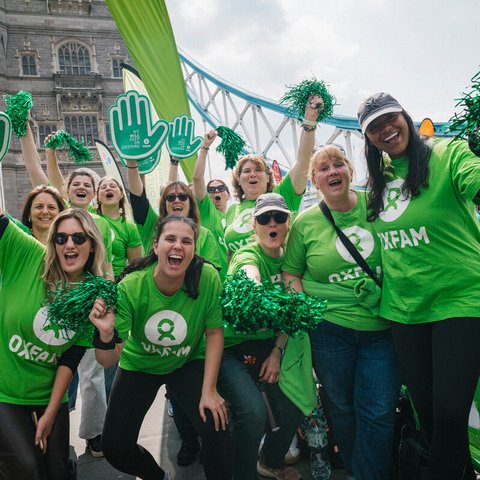 Oxfam volunteers, wearing green Oxfam logo t-shirts, pose for a group photo on Tower Bridge at the London Marathon 2025. They wave pom-poms and smile at the camera.