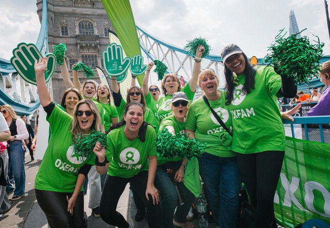 Oxfam volunteers, wearing green Oxfam logo t-shirts, pose for a group photo on Tower Bridge at the London Marathon 2025. They wave pom-poms and smile at the camera.