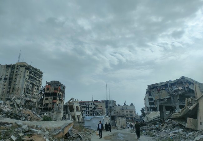 People walk in between destroyed buildings, turned to rubble
